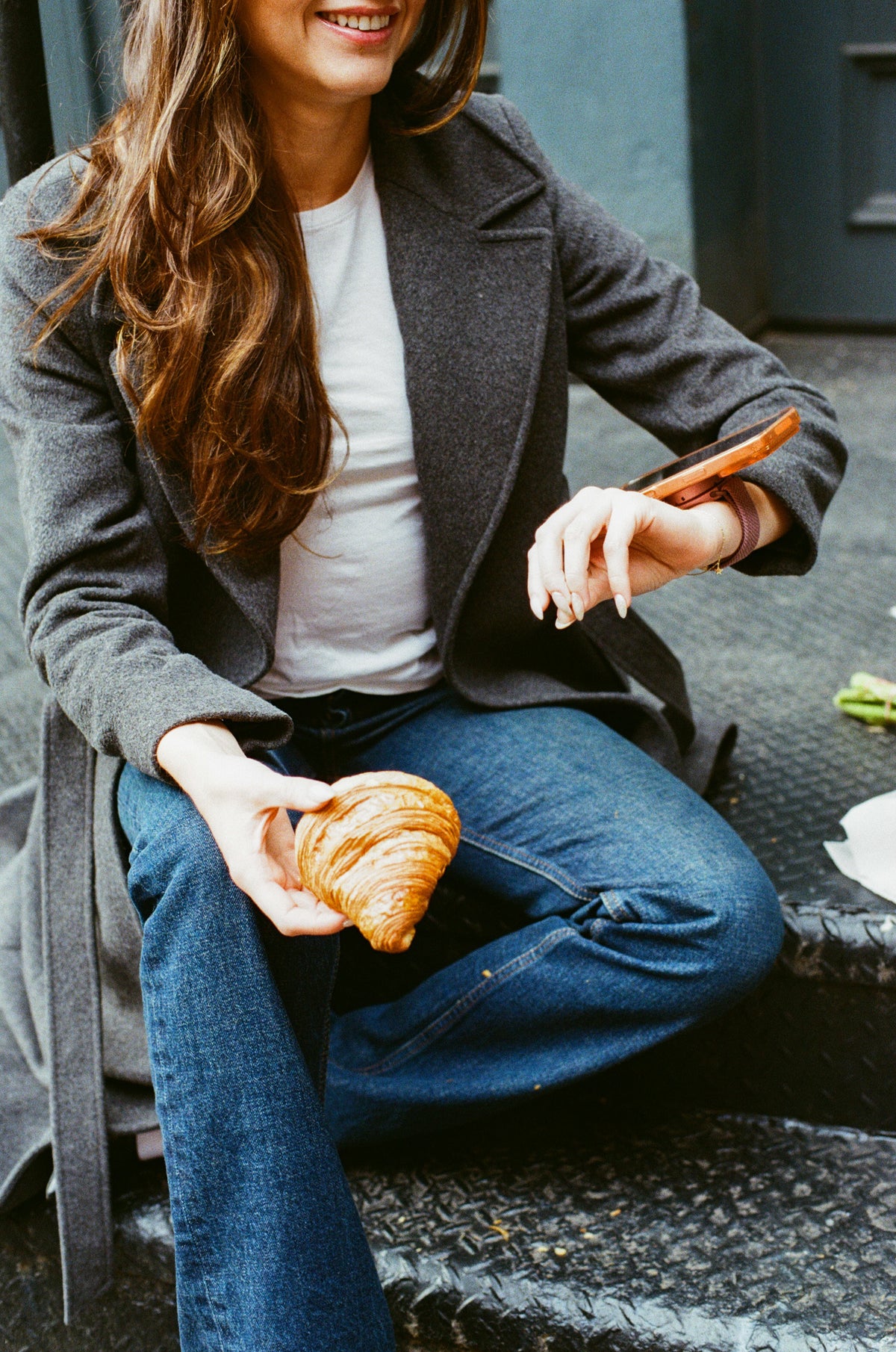 a woman sitting on a stoop with a croissant, phone on her wrist
